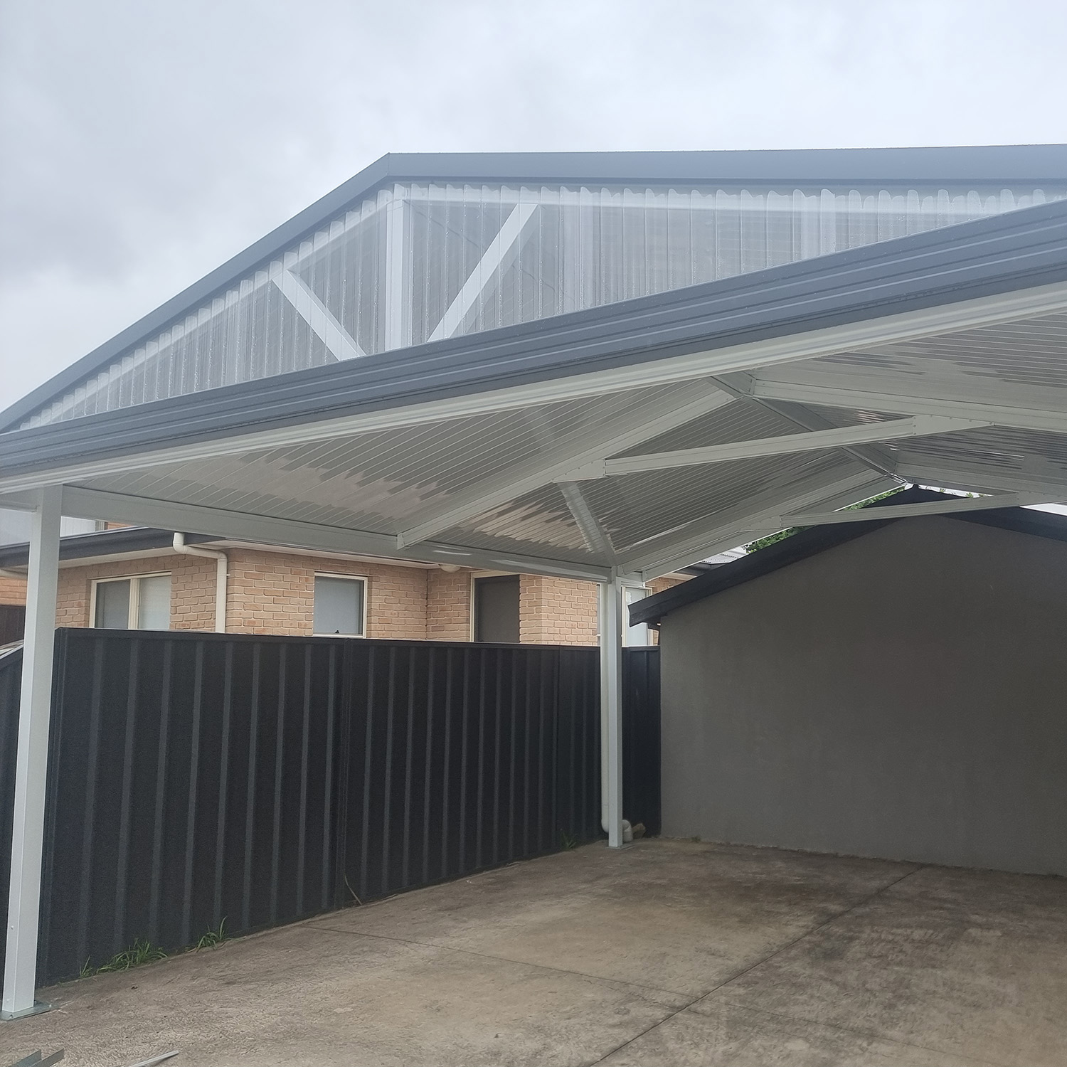 Modern DIY freestanding gable roof carport beside brick house and black fence.